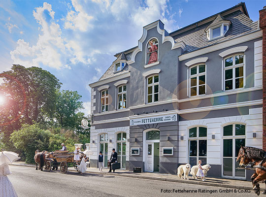 Eine strahlende Sonne beleuchtet ein Hotel. Menschen spazieren vorbei uns Kutschen werden auf der Straße vor dem Gebäude beladen.