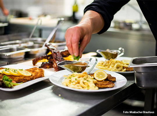 Blick "über die Schulter" von einem Koch, welcher Kräuter über ein Schnitzel mit Spätzle streut