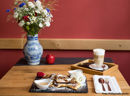 Anschaulich gedeckter Tisch mit Dessert, Kaffee, einem Apfel so wie ein Blumenstrauß